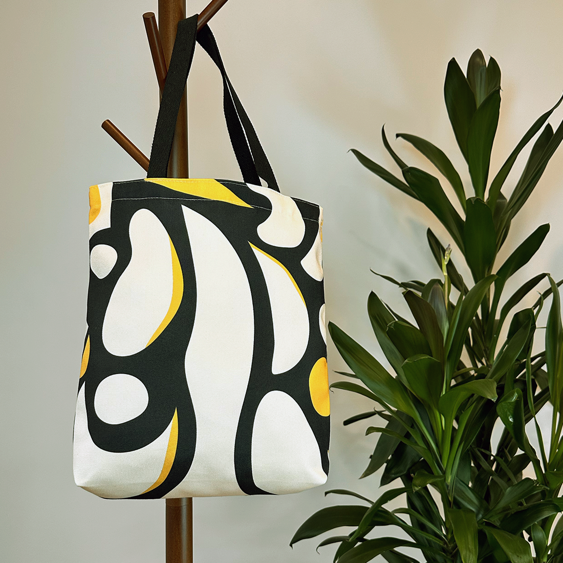 Abstract black and yellow tote bag on a wooden hanger next to a green plant, blending functionality and decor.