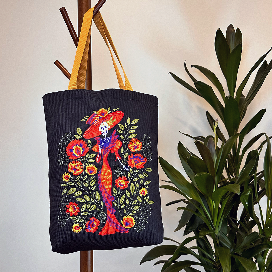 Lifestyle image of a black tote with a vibrant La Catrina skeleton and floral design, hanging on a coat rack with a yellow strap.