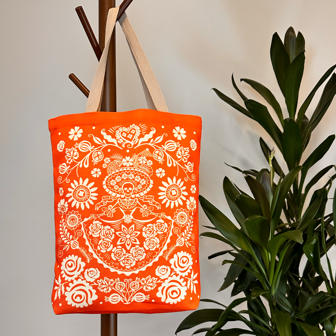 Orange tote bag with beige straps hanging on a coat rack. Highlights the vibrant white Day of the Dead pattern.