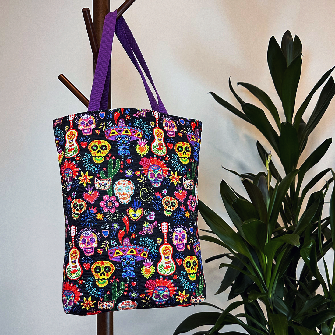 Lifestyle image of a black tote bag with bold Day of the Dead motifs and a purple strap, hanging on a coat rack.