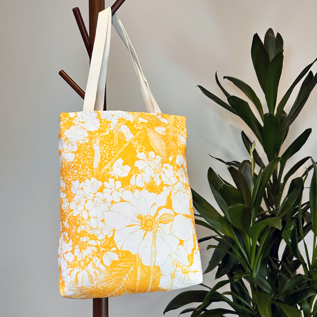 Lifestyle image of a yellow tote bag with white floral patterns, hanging on a coat rack. Adds a fresh and artistic touch.